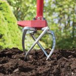 A red-handled manual garden cultivator with metal tines is digging into dark soil in a garden, ideal for package design or Advertising shoots, with green foliage and trees softly blurred in the background.